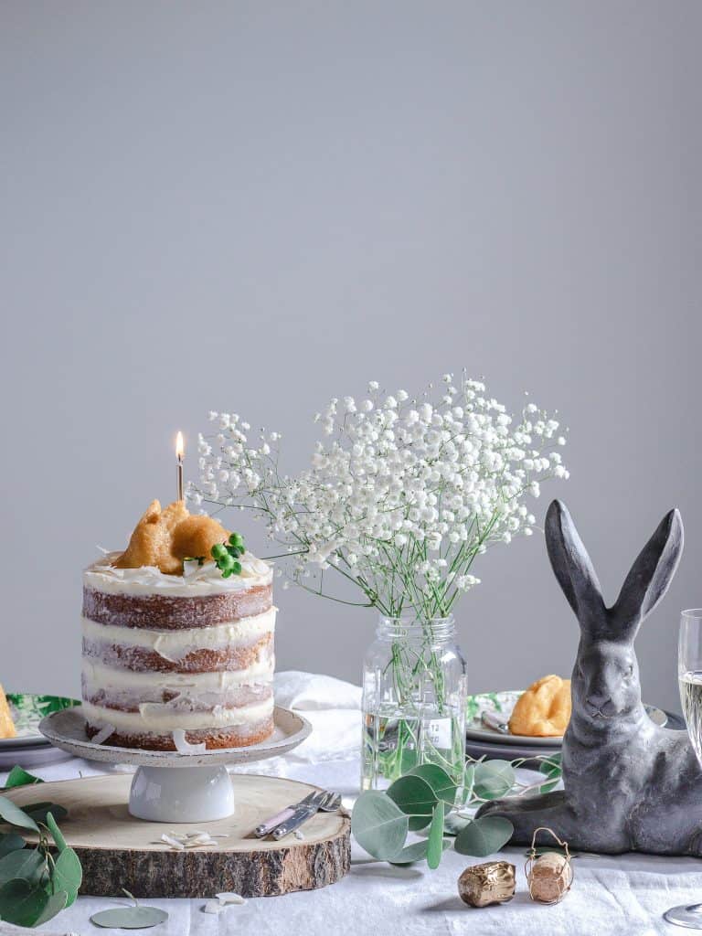 Tablescape with four layer lemon and coconut naked cake. Cake has a small bunny shaped cake on top with a lit gold candle. Tbale decorated with a Pottery Barn ceramic bunny