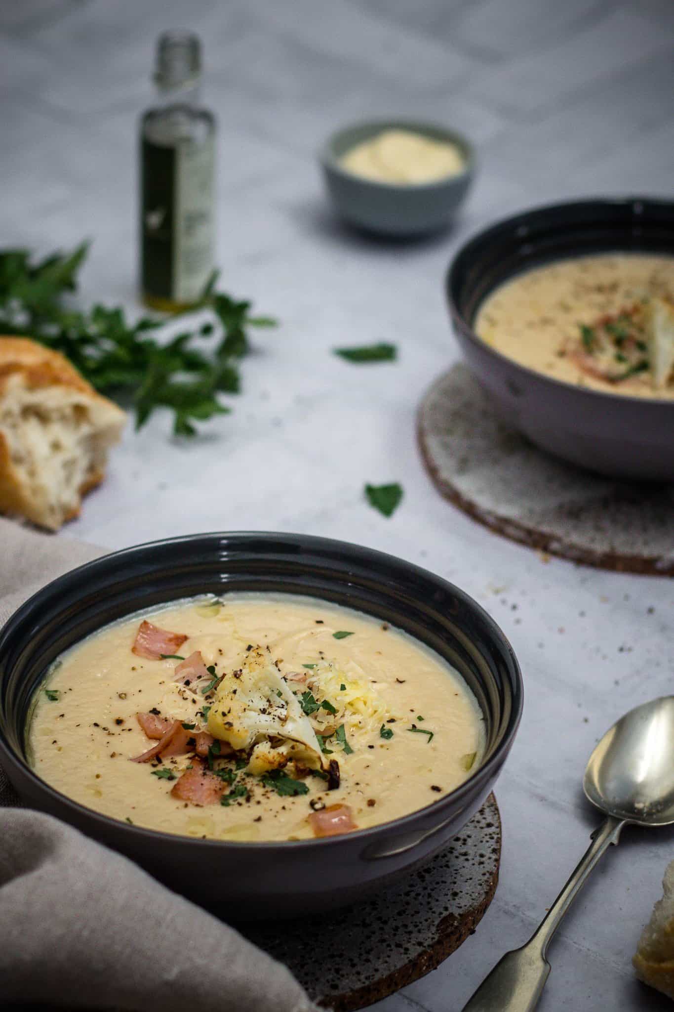 Two bowls of cheesy cauliflower and leek soup on a table along with a bottle of truffle oil and crusty brwad.