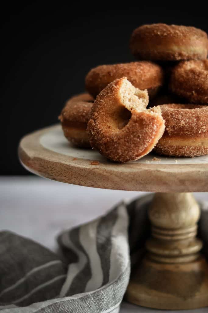 Timber and marble cake stand with doughnuts stacked on top. One doughnut has a bite taken out of it. 