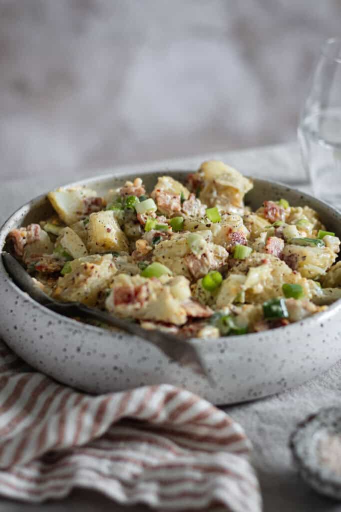 A bowl of egg and bacon potato salad on a linen tablecloth. A striped napkin, salt and a glass of water sit beside.