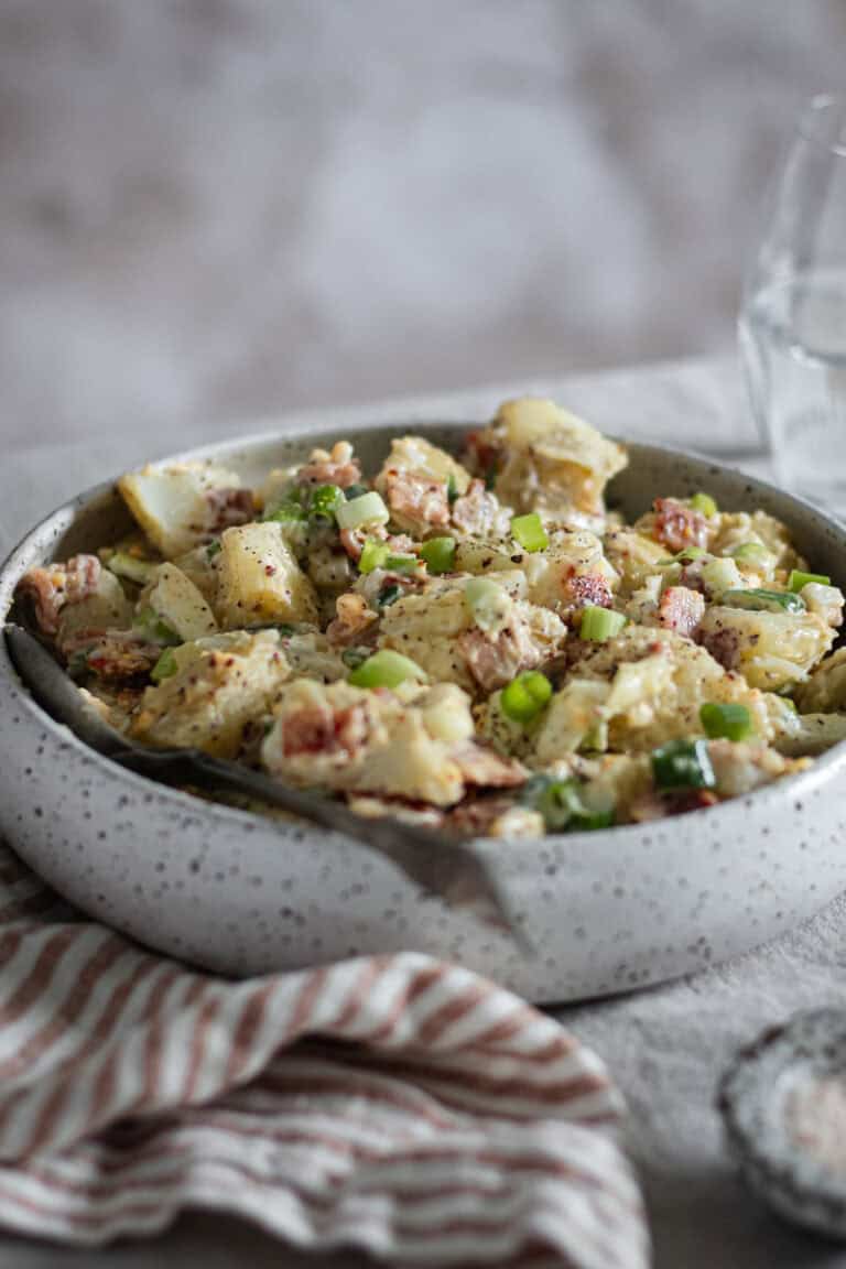 A bowl of egg and bacon potato salad on a linen tablecloth. A striped napkin, salt and a glass of water sit beside.