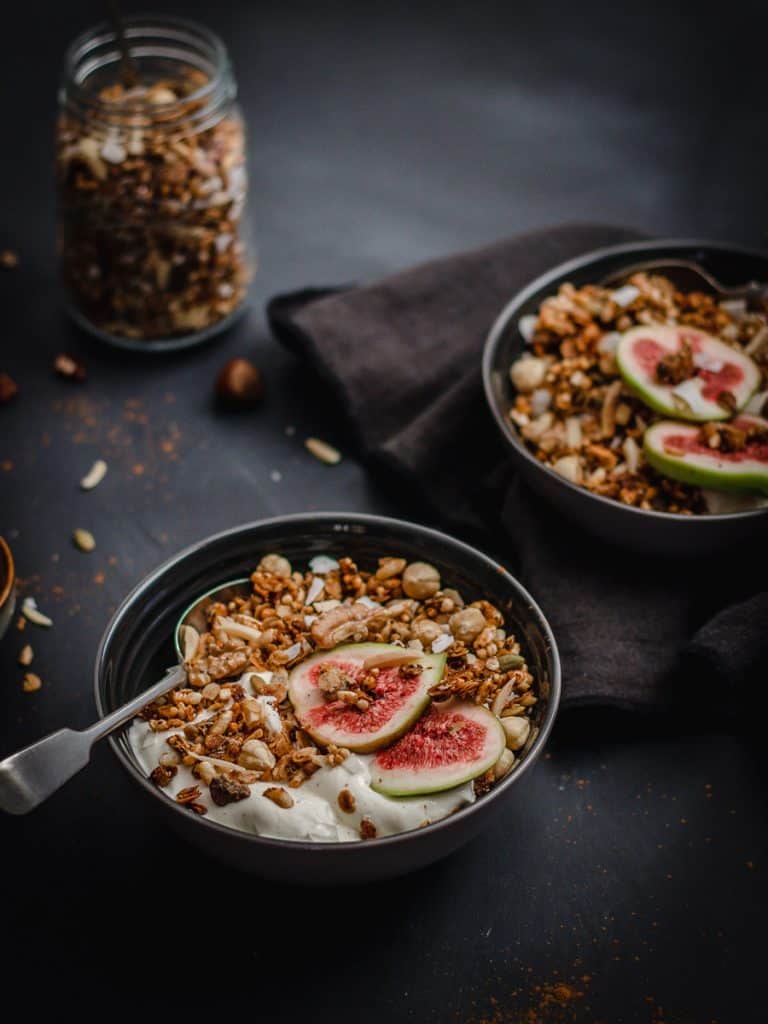 two grey bowls on a black background full of spiced honey nut granola, greek yoghurt and freshly sliced figs