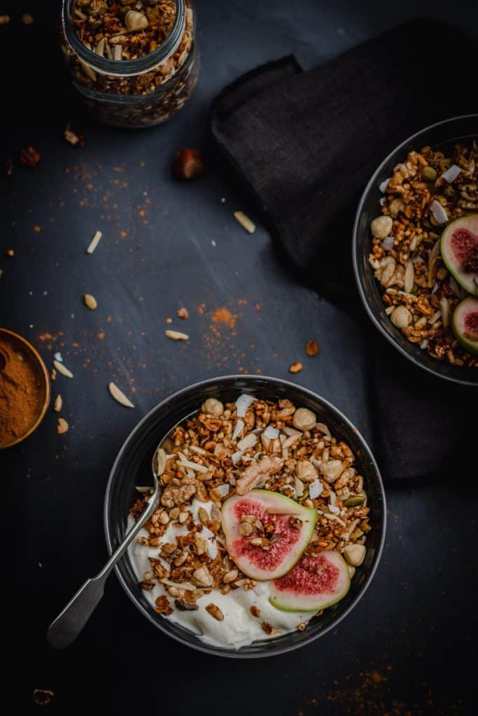 flatlay of two grey bowls on a black background full of spiced honey nut granola, greek yoghurt and freshly sliced figs. To the side is a small container of spice.