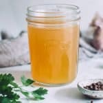 a jar of homemade chicken stock made in a pressure cooker. There is a bunch of parsley, some garlic and peppercorns on the bench around the jar