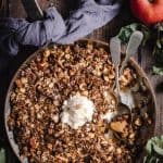 A Copper frypan of warm Apple and Walnut ANZAC Crumble. There is a scoop of ic cream melting on top and a spoonful of crumble eaten.