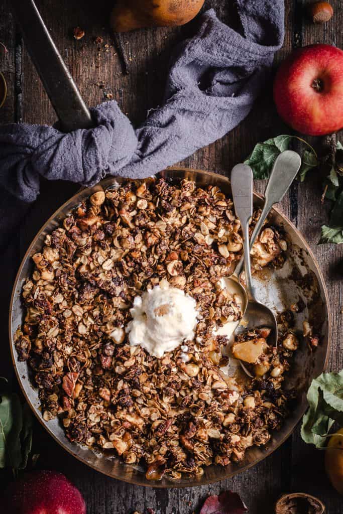 A Copper frypan of warm Apple and Walnut ANZAC Crumble. There is a scoop of ic cream melting on top and a spoonful of crumble eaten.