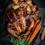 an oval roasting pan with a black garlic roast chicken, roast vegetables and peas in it.