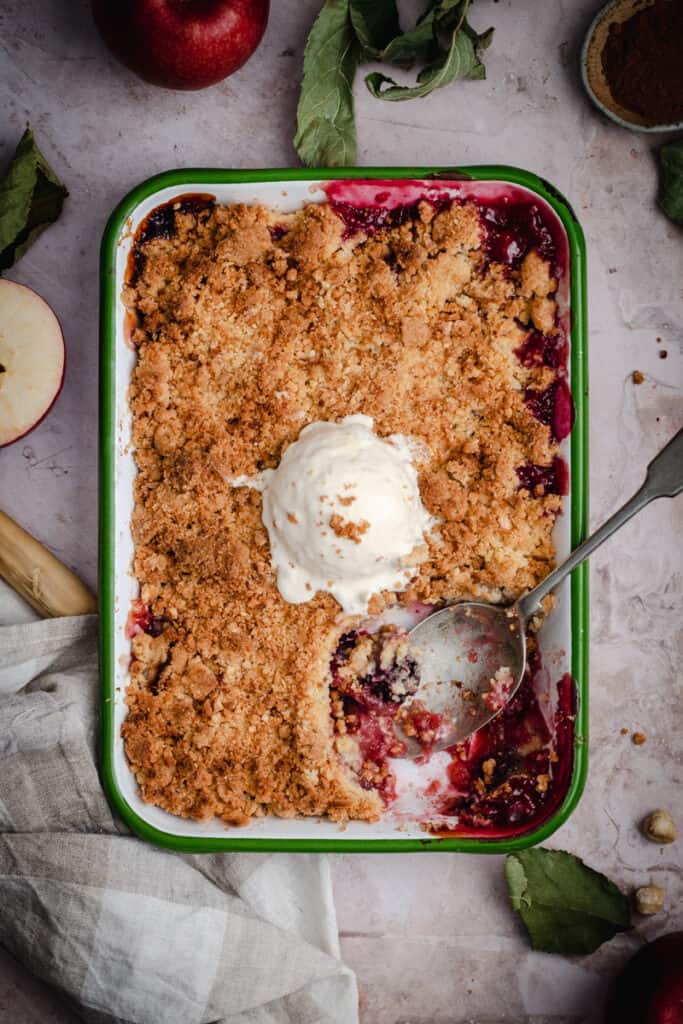 a flatlay of a apple and blackberry crumble in an enamel baking dish with ice cream on top. A scoop of crumble is eaten