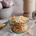 a jar of dehydrated garlic chips on a bench alongside a mortar and pestle and whole garlic bulbs