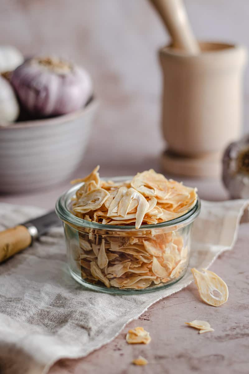 a jar of dehydrated garlic chips on a bench alongside a mortar and pestle and whole garlic bulbs