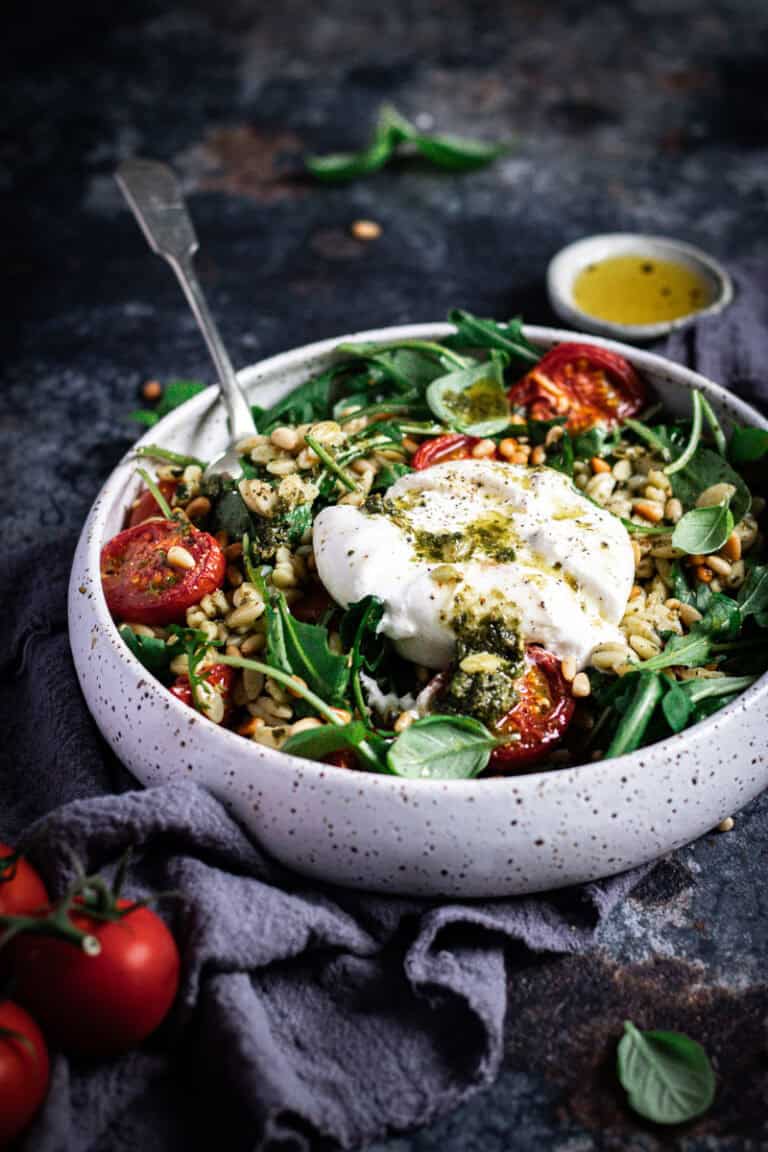 A large grey bowl of orzo pasta salad with a serving spoon in it. A split open burrata sits on top with pesto drizzled on top.