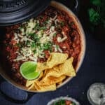 A black dutch oven full of black bean chili con carne with a chopped salad and corn chips.