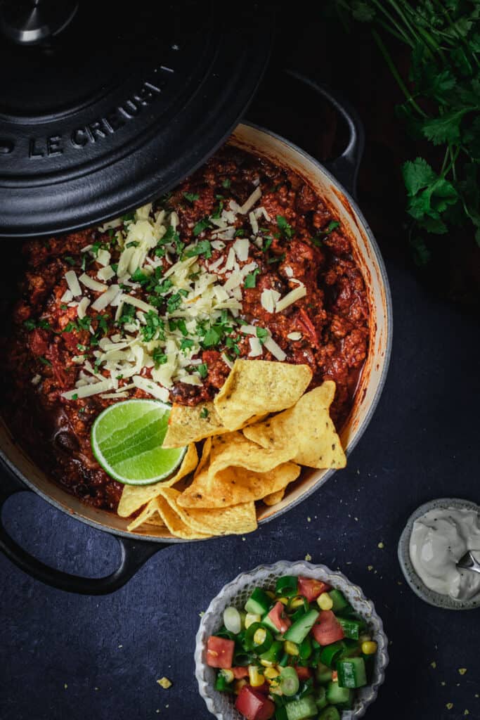 A black dutch oven full of black bean chili con carne with a chopped salad and corn chips.