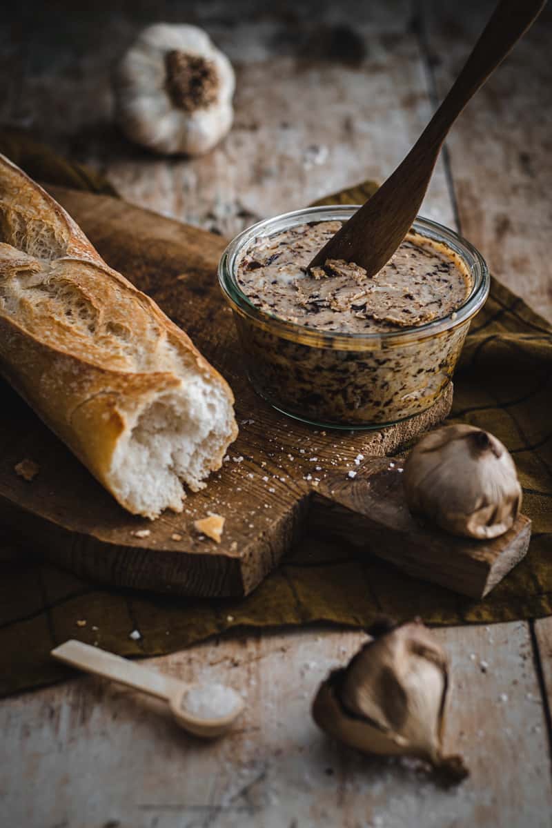 A jar of homemade black garlic butter on a board alongside a chunk of crusty baguette.