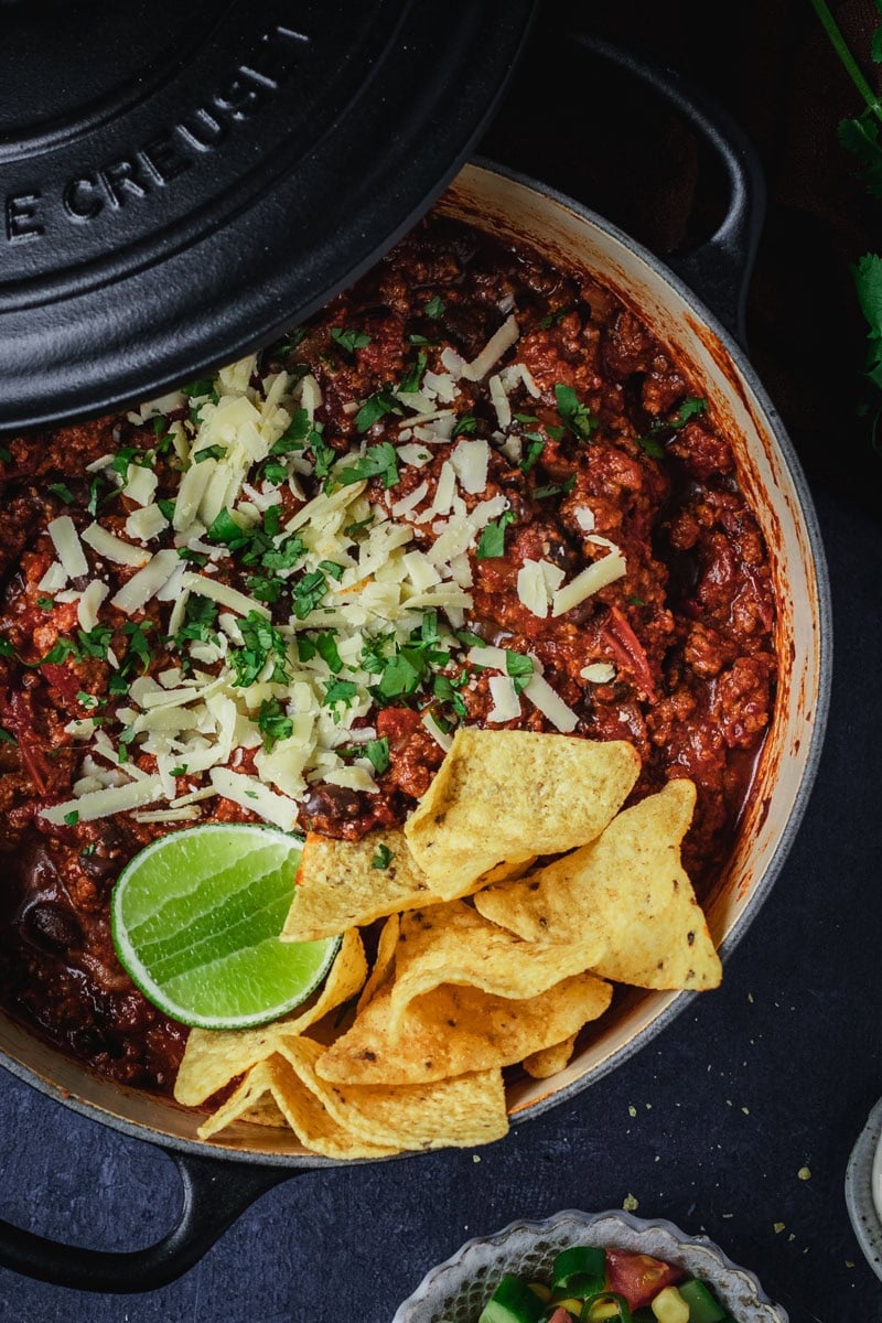 A dutch oven full of chili con carne. A few corn chips and a wedge of lime sit on one side.