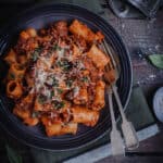 A bowl of slow cooked rigatoni bolognese on a table alongside a glass of wine.