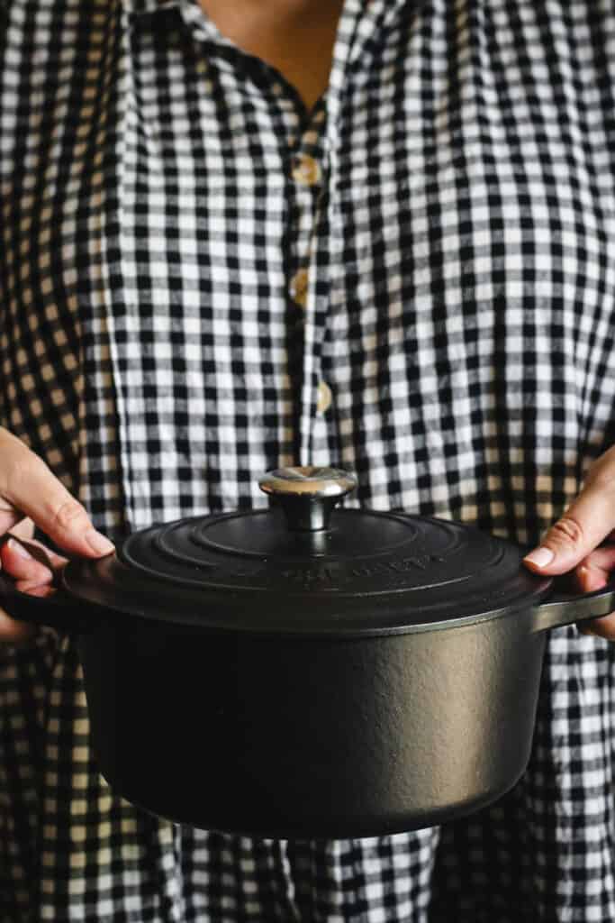 A woman wearing a black and white check dress holding a black Le Creuset enameled Dutch oven.