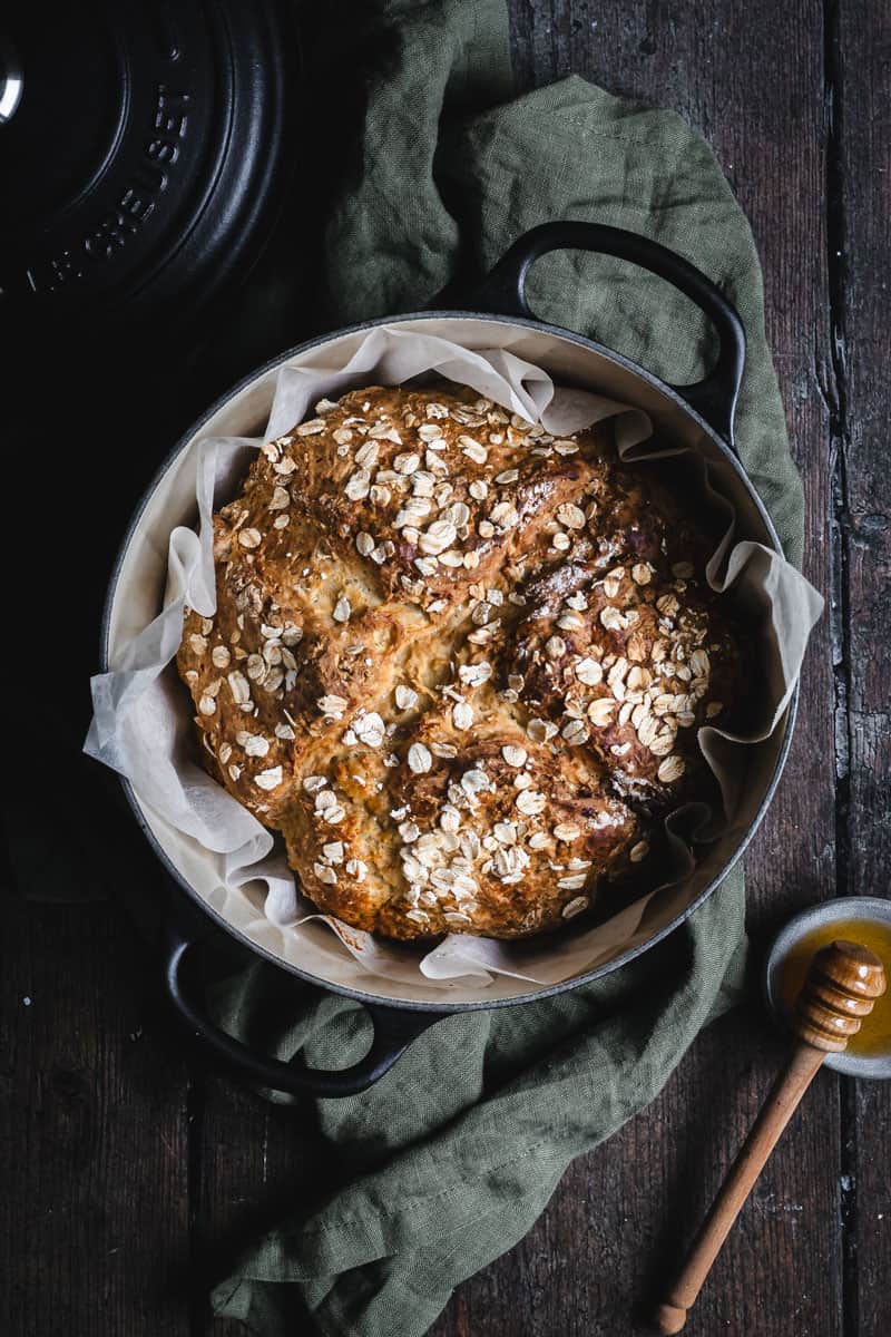 A black cast iron casserole with a golden loaf of soda bread in it. A small bowl of honey with a honey dipper sit beside.