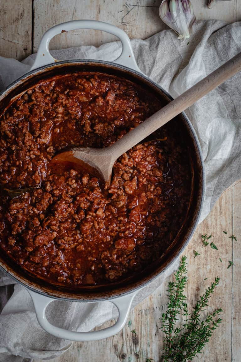 A white cast iron pot with slow-cooked Italian Pork and Nduja Rugu in it. A wooden spoon is stirring the Ragu.