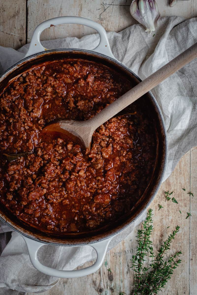 A white cast iron pot with slow-cooked Italian Pork and Nduja Rugu in it. A wooden spoon is stirring the Ragu.