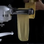 A woman feeding a sheet of homemade pasta through a KitchenAid pasta maker.