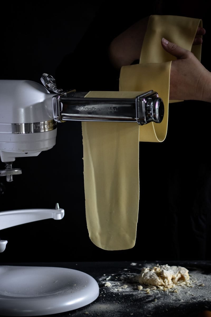 A woman feeding a sheet of homemade pasta through a KitchenAid pasta maker.