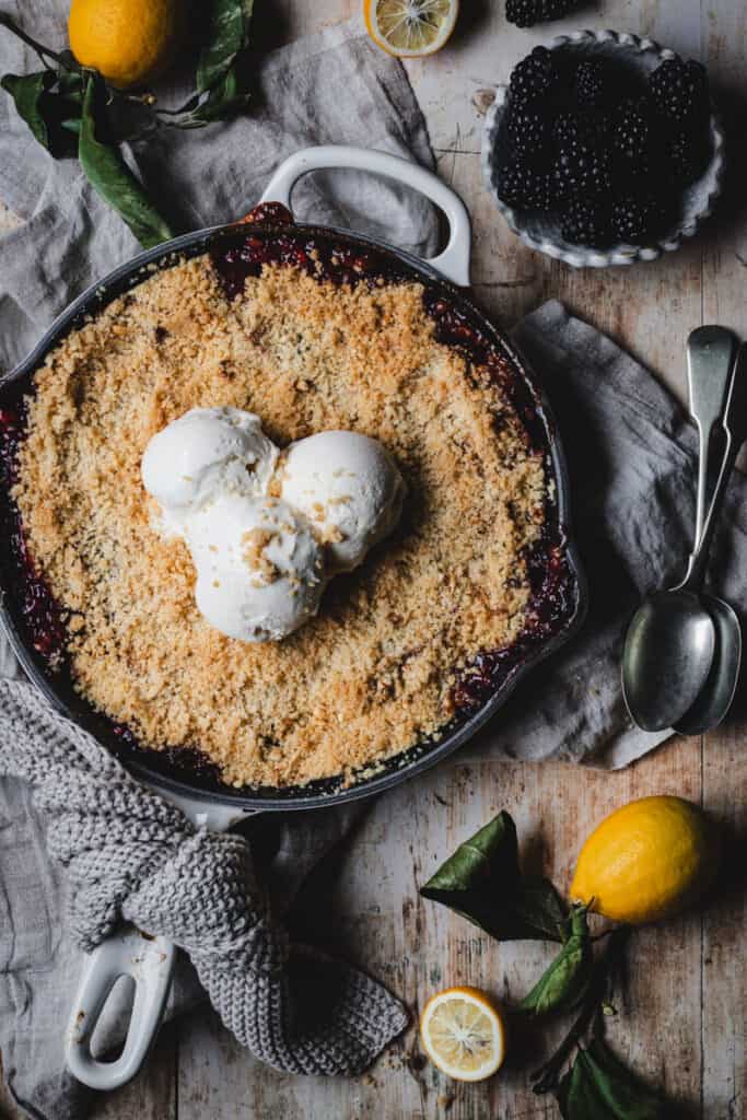 The finished Apple and Blackberry Crumble in a cast iron skillet. The dish sits on a kitchen bench surrounded by lemons, a bowl of blackberries and two silver spoons.