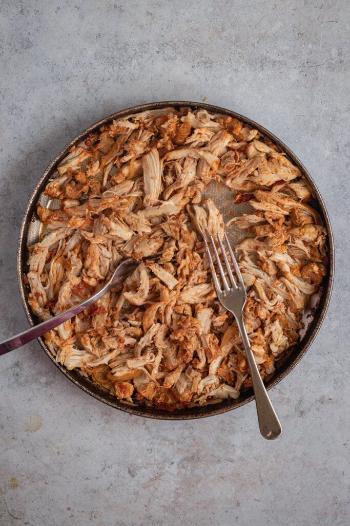 Two forks shredding the chicken thighs on a round metal dish.