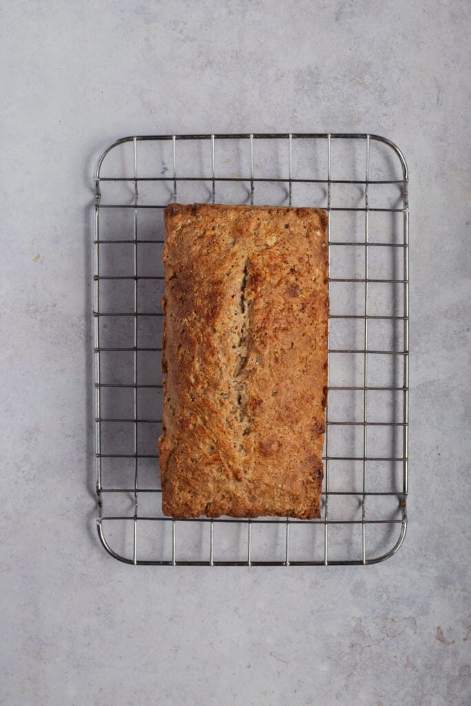 The baked loaf of brown bread cooling on a wire rack.