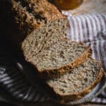 A loaf of Irish brown bread with a few slices cut. It sits on a stripe tea towel alongside a small knife and pot of honey.