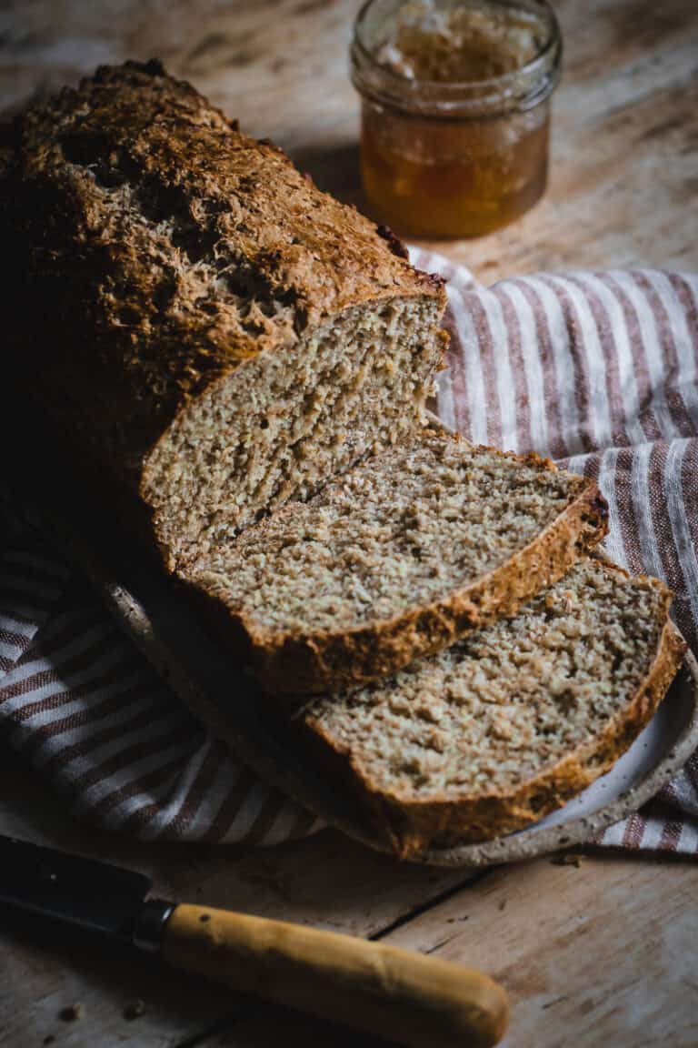 A loaf of Irish brown bread with a few slices cut. It sits on a stripe tea towel alongside a small knife and pot of honey.