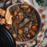 A black cast iron Dutch oven of beef and Guinness stew. A Couple of pieces of bread are sitting in the side of the pot and fresh rosemary sits in a small bowl to one side.