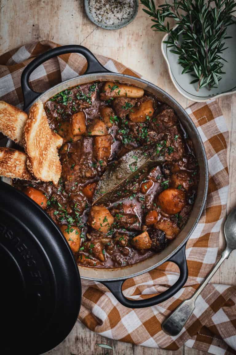 A black cast iron Dutch oven of beef and Guinness stew. A Couple of pieces of bread are sitting in the side of the pot and fresh rosemary sits in a small bowl to one side.
