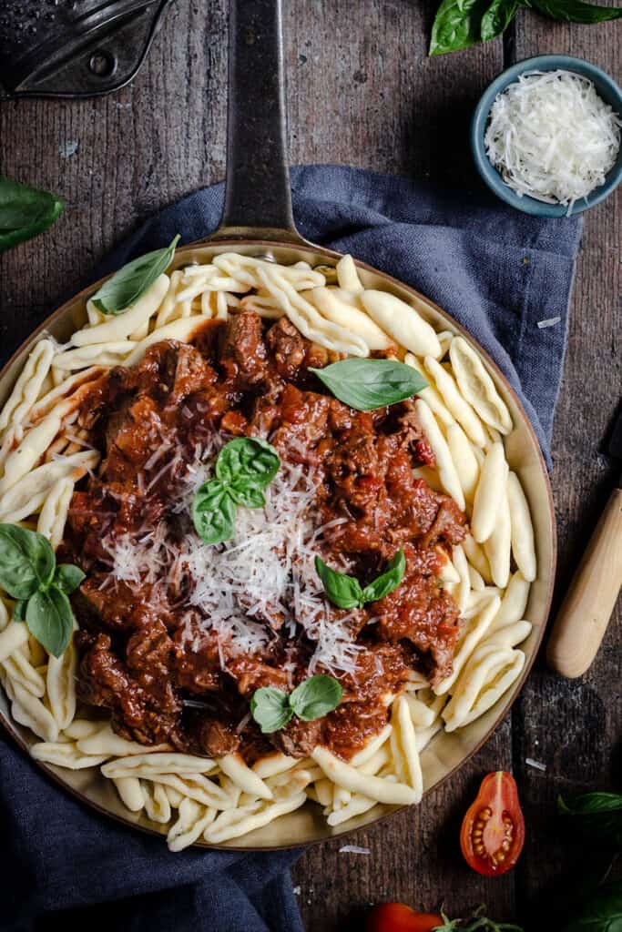 flatlay of a frypan of slow cooked beef and red wine ragu on a bed of capunti pasta, with parmasean and basil leaves.