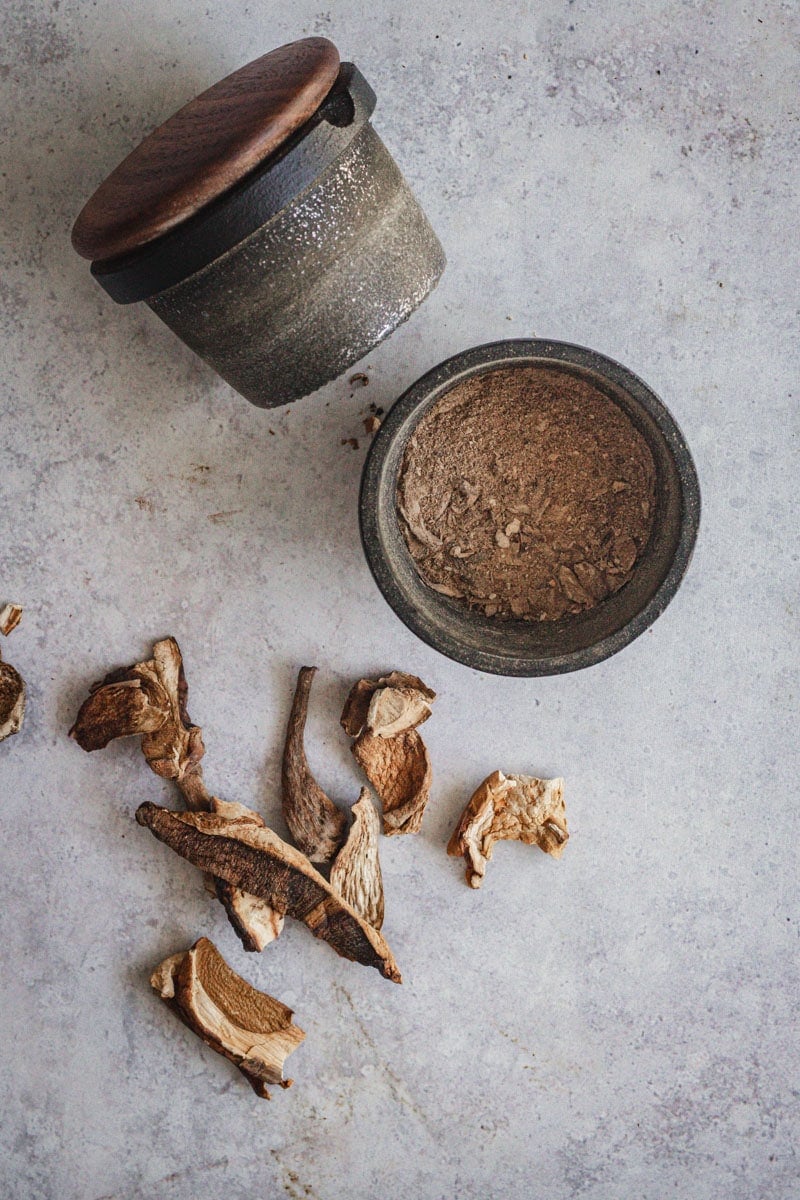 A cast iron spice grinder with ground up porcini slices in it. Some whole porcini slices sit beside.