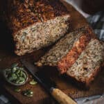 A loaf of porridge bread with two slices cut sits on a wooden board. A small knife and a bowl of pepitas sit beside on the board.
