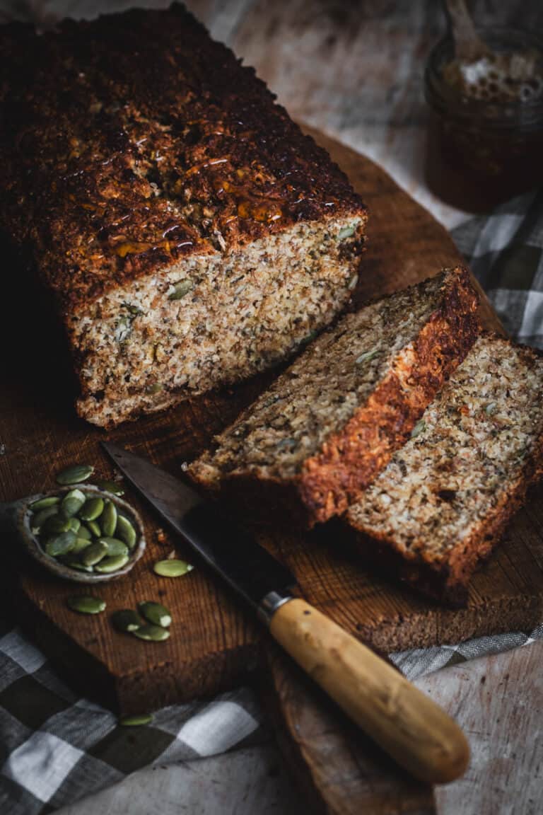 A loaf of porridge bread with two slices cut sits on a wooden board. A small knife and a bowl of pepitas sit beside on the board.