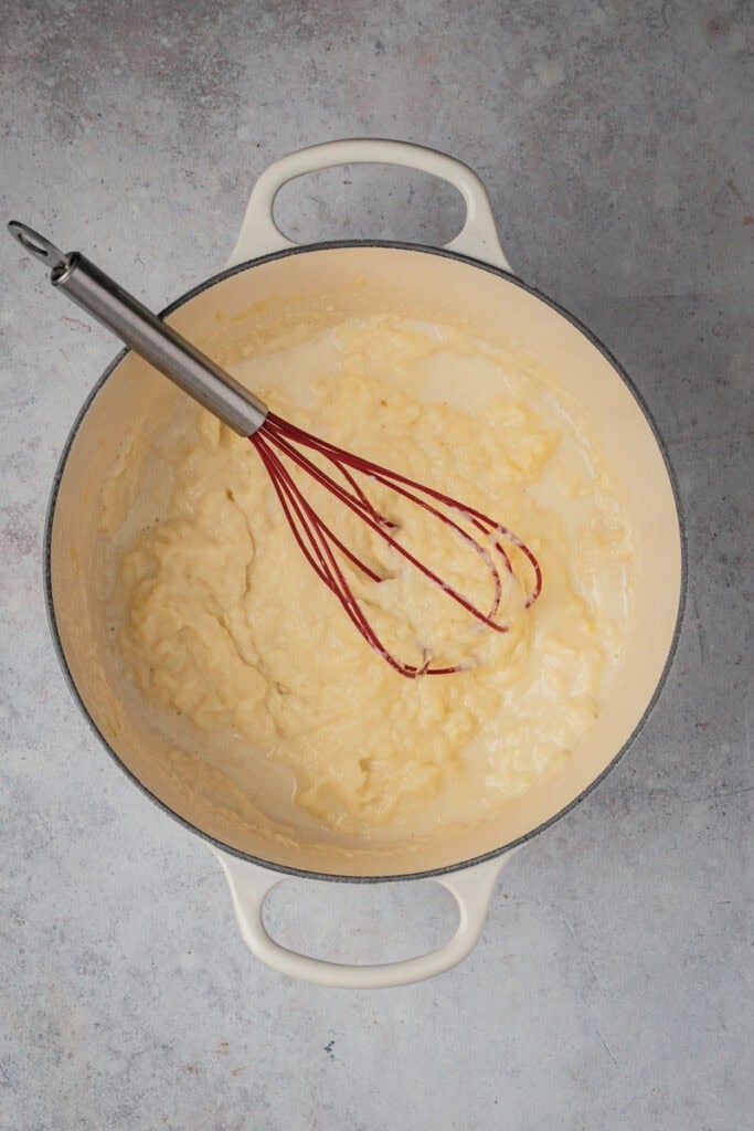 Milk has been added to the pot and the sauce is being mixed with a red whisk.