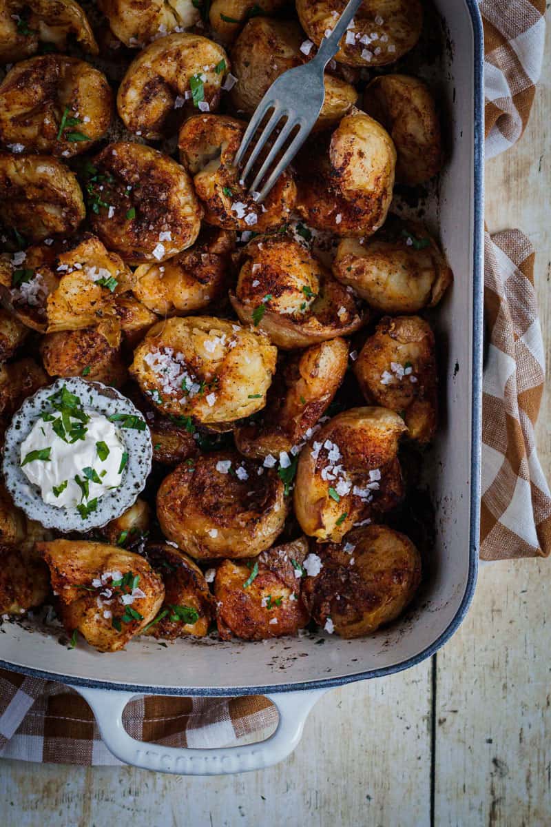 A close up of a cream enameled cast iron pan full of cajun spiced roast potatoes. A small bowl of sour cream sits on top.
