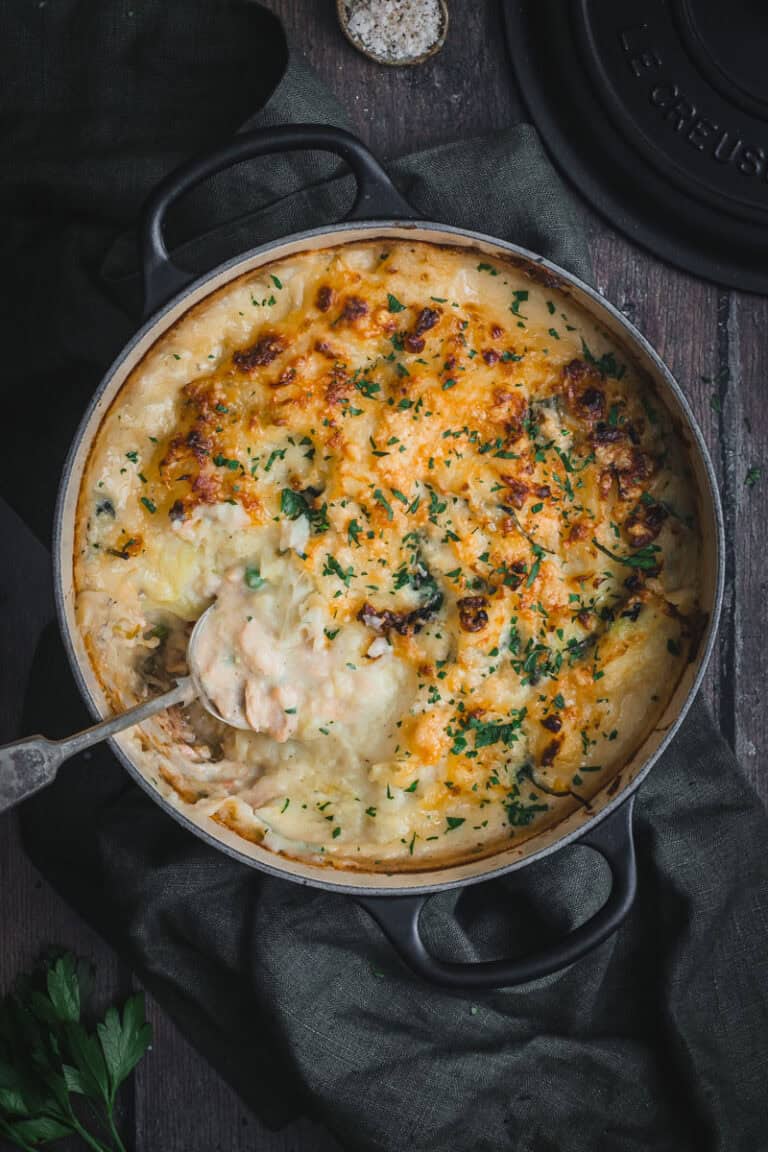 A black Le Creuset cast iron Dutch oven with a fish pie in it. A serving has been scooped out revealing the creamy seafood mornay inside.