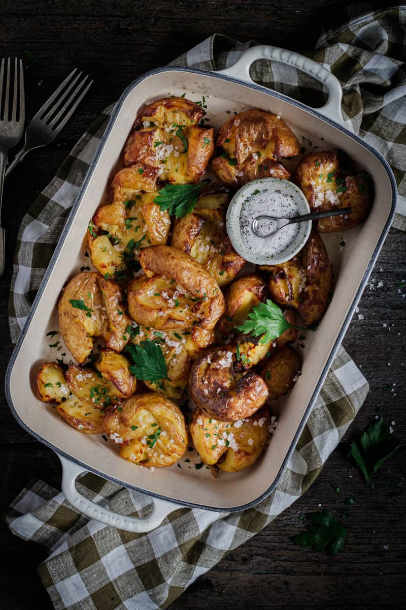 A white enamel cast iron roasting pan full of smashed duck fat roast potatoes. A bowl of truffle salt sits on top.