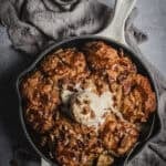 A apple cobbler in a cream Le Creuset skillet drizzled with butterscotch sauce and a scoop of ice cream.