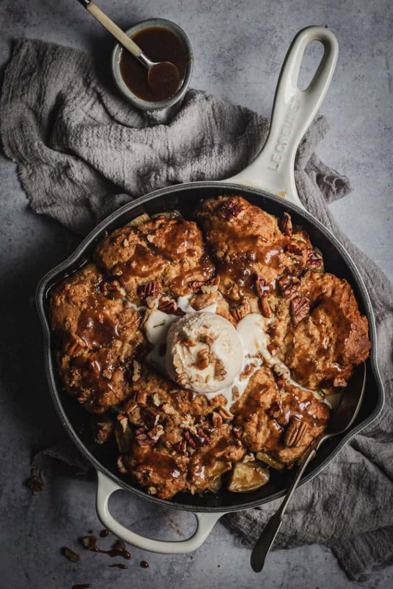 A apple cobbler in a cream Le Creuset skillet drizzled with butterscotch sauce and a scoop of ice cream.