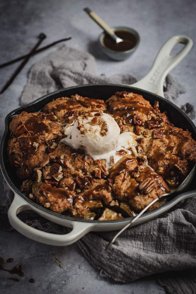 A cooked apple cobbler in a cream colored skillet. A large scoop of icecream is on top and a spoon sits in the side.