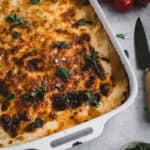 A large white enamel dish of ragu lasagna with sage leaves scattered on top. A knife and truss tomatoes sit beside.