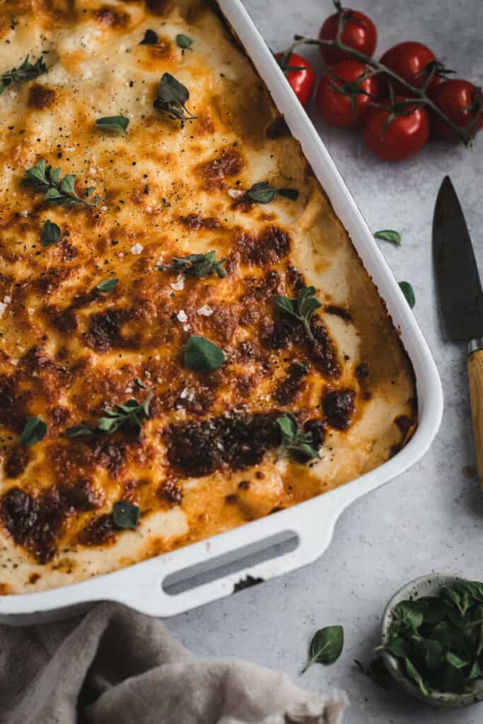 A large white enamel dish of ragu lasagna with sage leaves scattered on top. A knife and truss tomatoes sit beside.