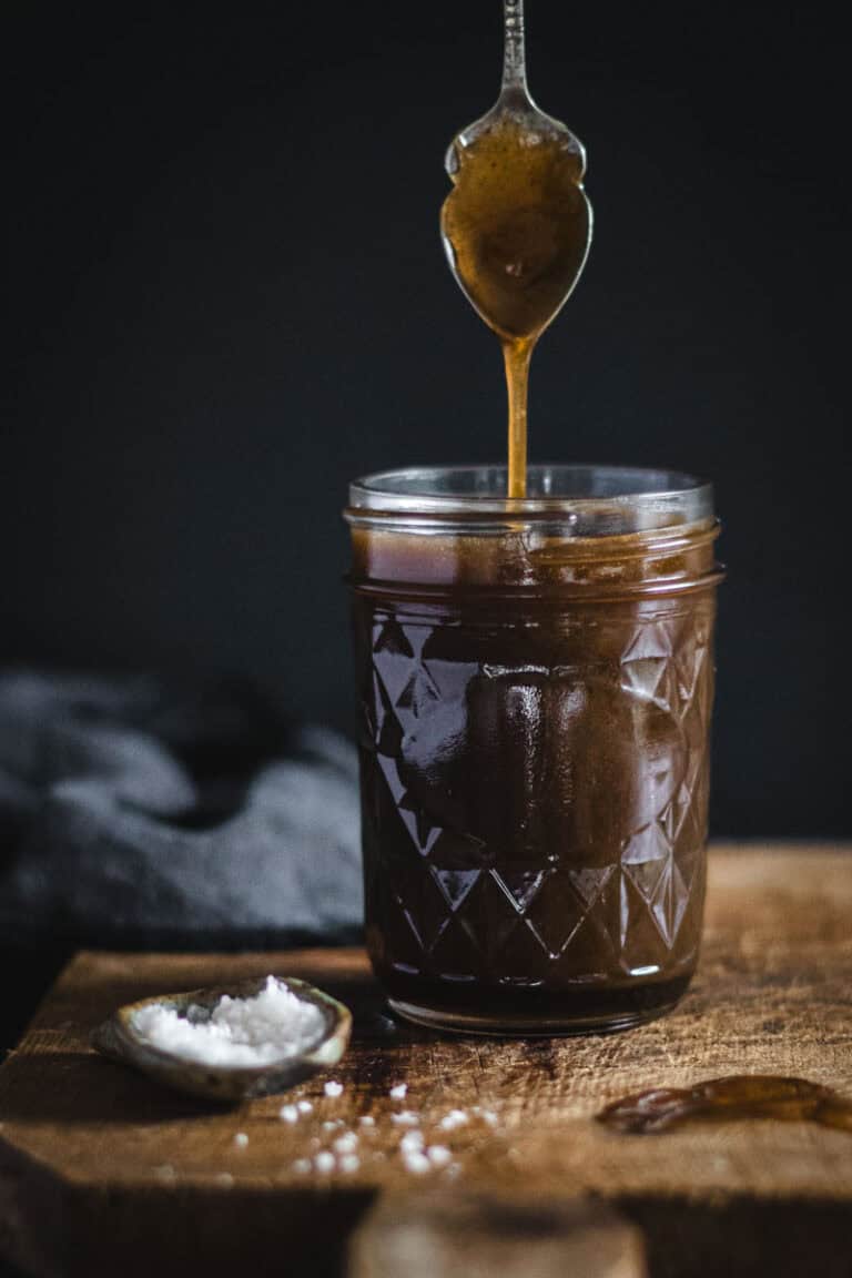 A jar of whisky caramel sauce on a timber board. A spoon has been dipped in the sauce from above.
