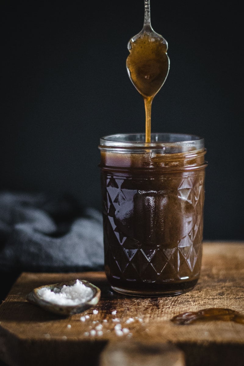 A jar of whisky caramel sauce on a timber board. A spoon has been dipped in the sauce from above.