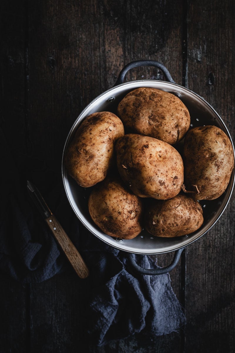 A white colander full of large, dirt covered sebago potatoes. A timber peeler sits beside.