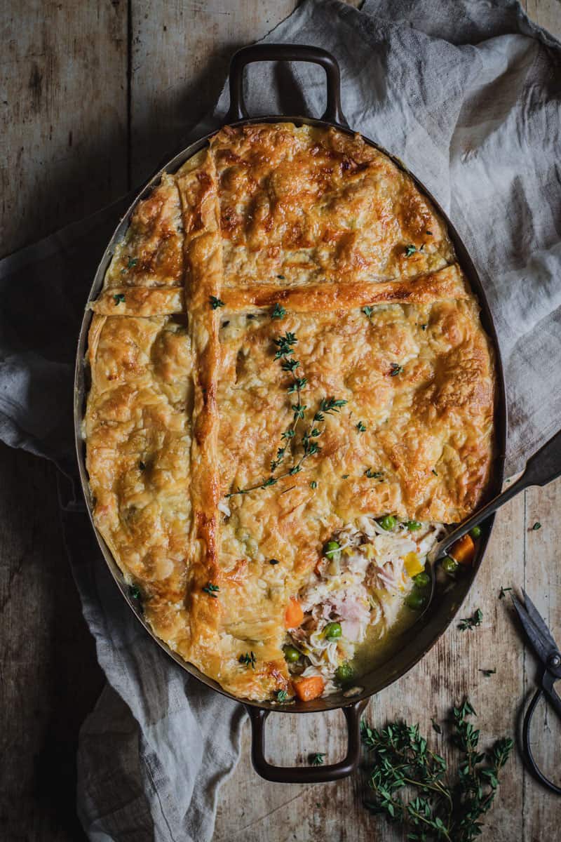 An oval pan full of chicken bacon pie topped with golden puff pastry. Fresh thyme and small back scissors sit beside it.